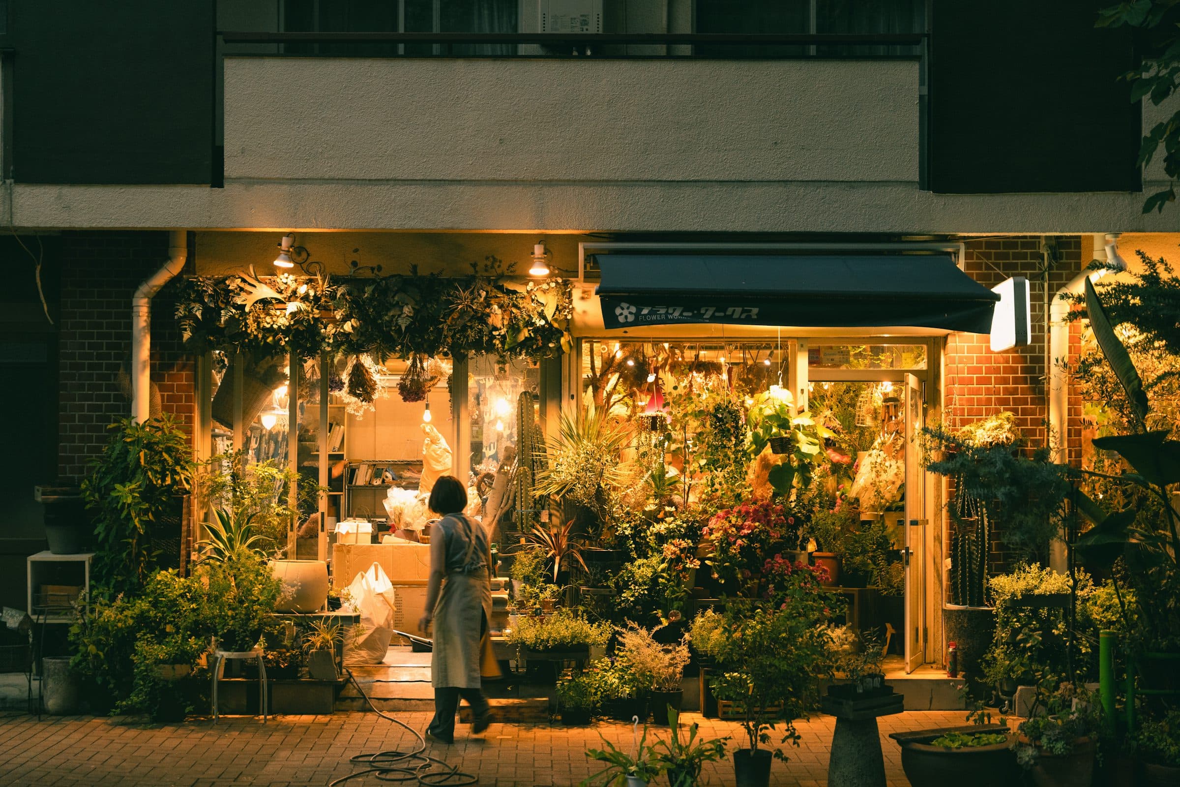 A florist walks past a flower shop glowing warmly late at night.